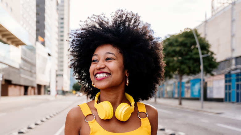 Woman with curly afro smiling on street in yellow top and yellow headphones around her neck