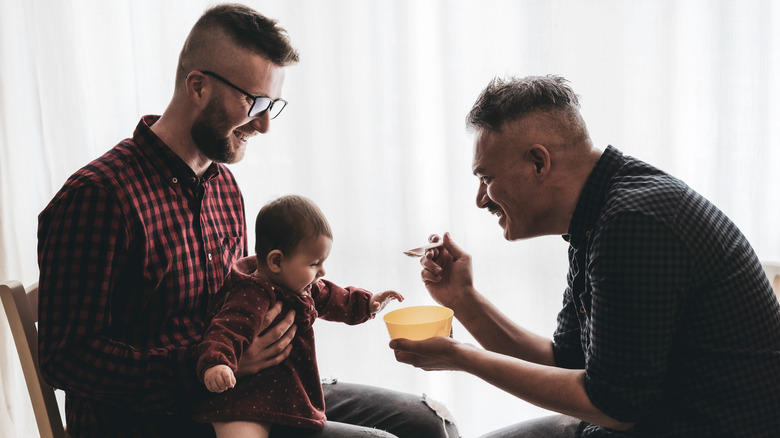 Baby on man's lap while second man feeds baby with food on spoon