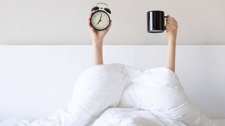 Woman in bed holding up an alarm clock and mug