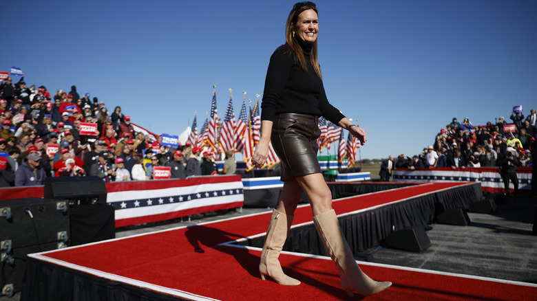 Sarah Huckabee Sanders walking on a red carpet outside at a Trump rally; she's wearing a black turtleneck, leather skirt, and suede boots.