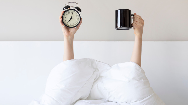 Woman hands coming out from comforter and holding alarm clock and coffee mug