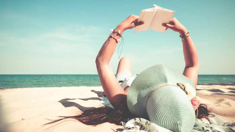 woman laying on the beach holding up a book