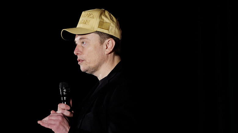 Elon Musk wearing a gold MAGA hat and holding a microphone