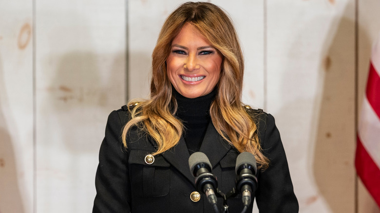 Melania Trump grinning behind a microphone, wearing a brass-button blazer.