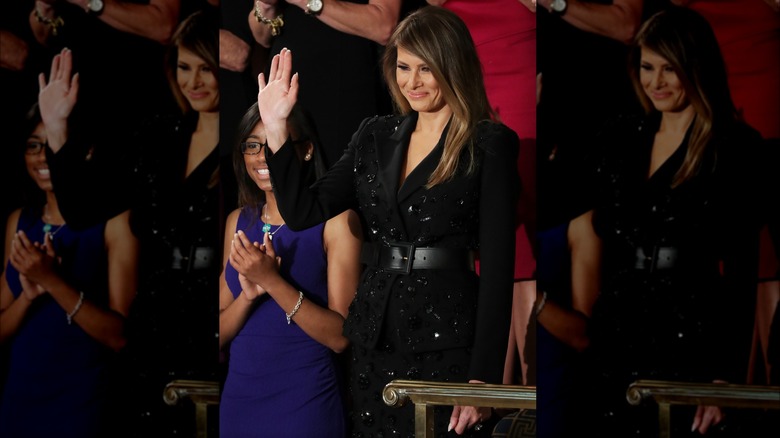 Melania Trump wearing a sparkly black outfit, closed smile, waving to a crowd in congress.