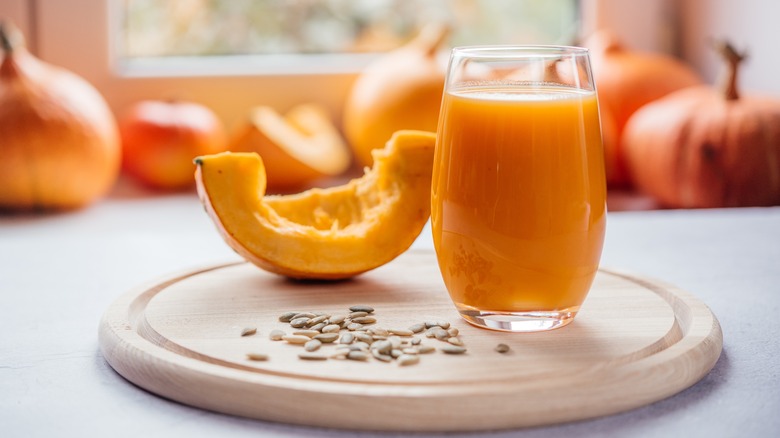 A glass of pumpkin juice, a slice of pumpkin, and pumpkin seeds on a counter