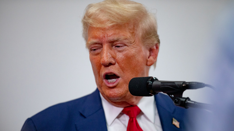 Donald Trump in the middle of speaking, with a very red face, red tie, and yellow hair.