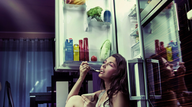 woman sitting in front of open fridge eating