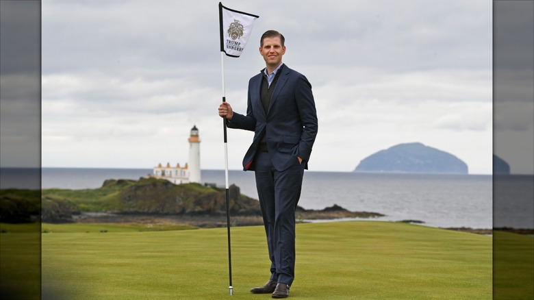 Eric Trump stands on a golf course, holding a golf flag