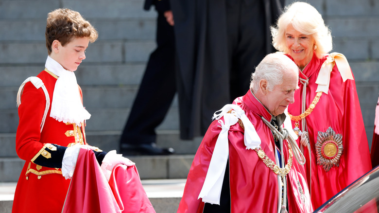 Lord Oliver Cholmondeley holding King Charles' robe with Queen Camilla smiling