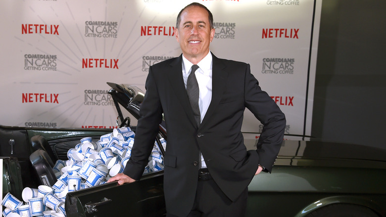 Jerry Seinfeld smiling and standing in front of a car