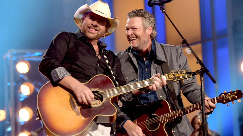 Toby Keith smiling and playing guitar, Blake Shelton smiling at Keith playing guitar