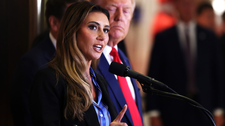 Alina Habba speaking at a microphone with Donald Trump frowning behind her