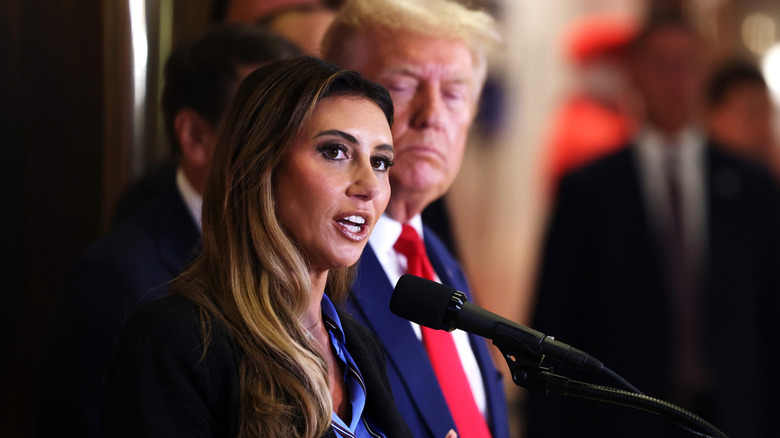 Alina Habba speaking into a microphone, with Donald Trump in the background.