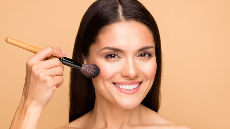 Brunette woman smiling and applying foundation with a brush.