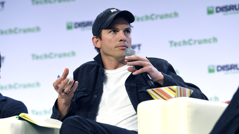 Ashton Kutcher sitting in a chair on stage with a microphone