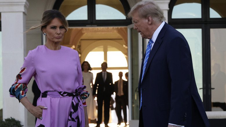 Melania Trump purple jumpsuit hand on hip with Donald