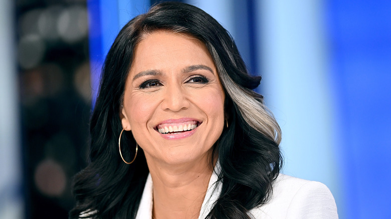 Tulsi Gabbard smiling in white blazer against blurred background