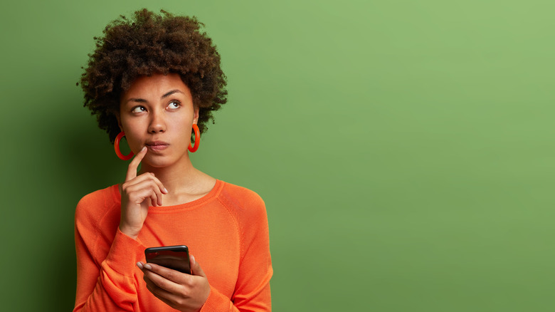 Woman with short brown curly hair wearing bright orange hoop earrings and a bright orange top puts her finger to her mouth and looks up at the sky quizzically while holding a cell phone in her other hand