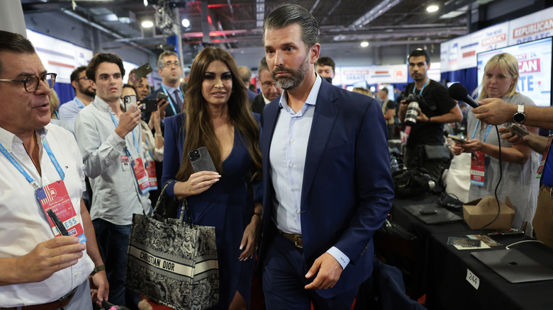 Kimberly Guilfoyle and Donald Trump Jr. walking through crowd of reporters holding phones and mics