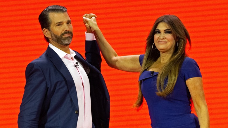 Donald Trump Jr. and Kimberly Guilfoyle holding hands on stage at an event.