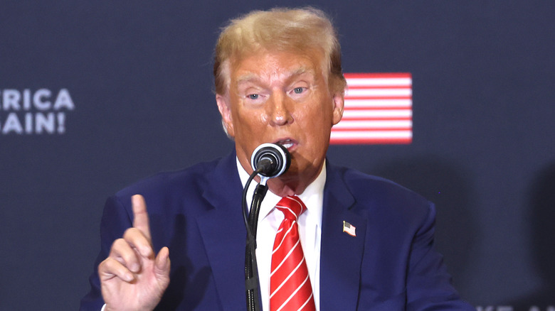 Donald Trump raises a finger as he speaks on a microphone with a bad tan