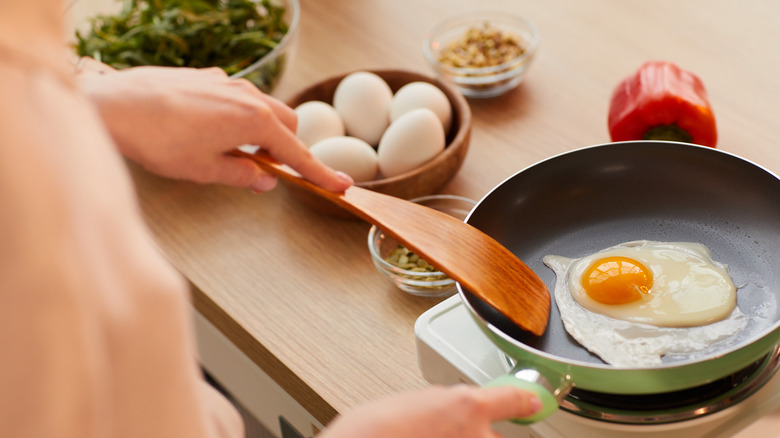 Woman frying egg on pan with bowls of leafy greens, eggs, and red bell pepper next to pan