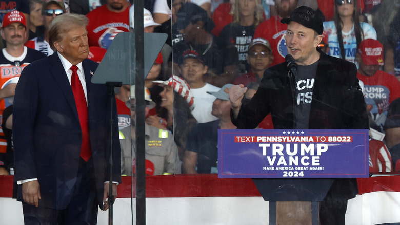 Elon Musk speaking at Trump podium while Donald Trump watches