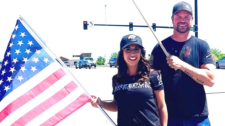 Lauren and Jayson Boebert holding American flags at an intersection, both in black T-shirts and smiling.