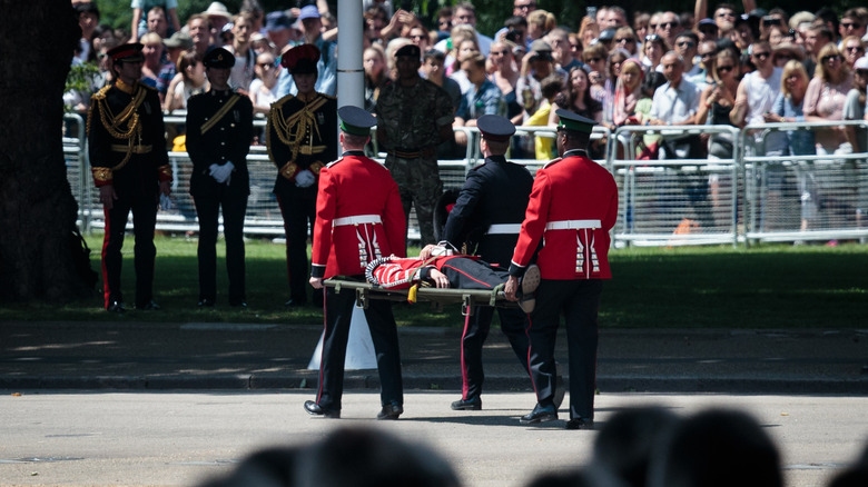 Royal guard being carried on a stretcher by other guards as the public watches on