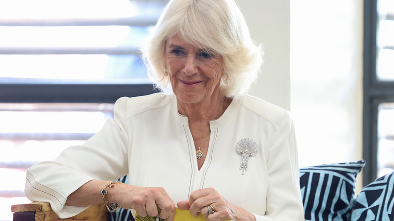 Camilla smiles, holds book on lap, jewelry on wrists and fingers visible