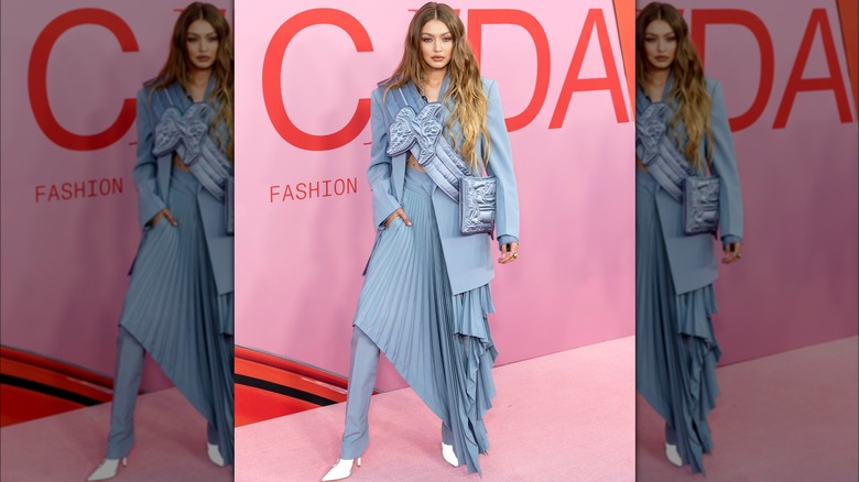 Gigi Hadid on the red carpet wearing a bulky, multi-layered outfit in blue, including pants, skirt, jacket, and sash.