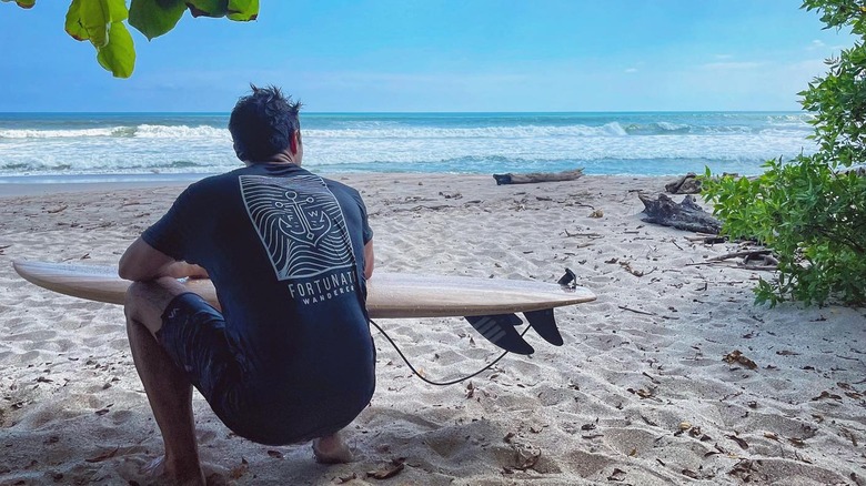 Ryan Paevey squatting on the beach with a surfboard