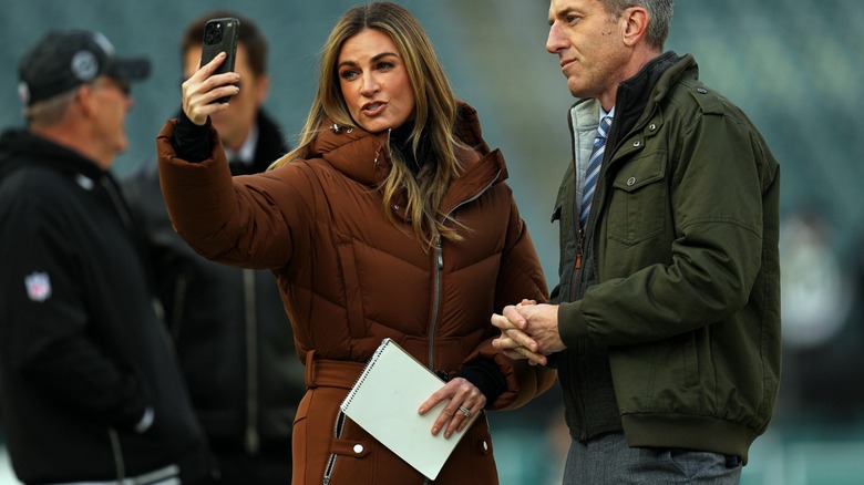 Erin Andrews taking a video with her phone, dressed in a brown puffy ski suit.