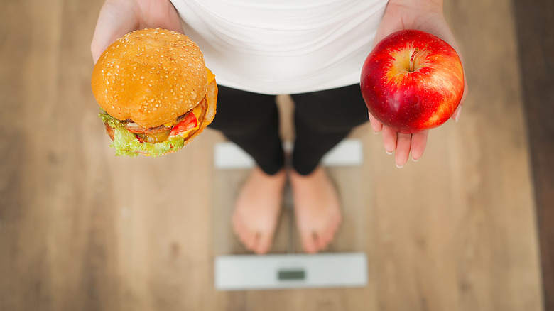 woman on scale with cheeseburger in one hand and apple in the other