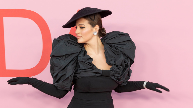 Ashley Graham wears a black hat, dress, and gloves.