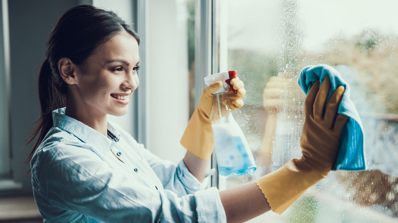 Young woman wearing yellow rubber gloves cleans window with spray bottle