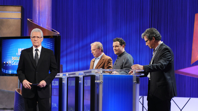 Jeopardy! host Alex Trebek stands in front of three contestants
