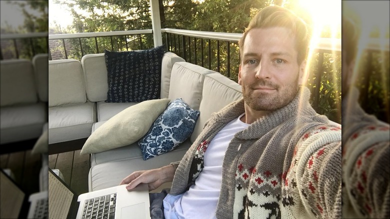 Orlando Soria sitting on an outside sofa, sun glinting over his shoulder, laptop open, and wearing a cardigan sweater.