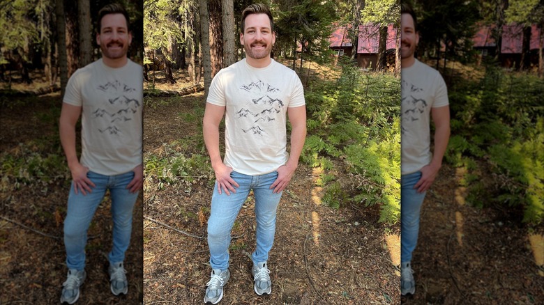 Orlando Soria in the woods, wearing a graphic t-shirt, blue jeans, and sneakers, smiling under his mustache.