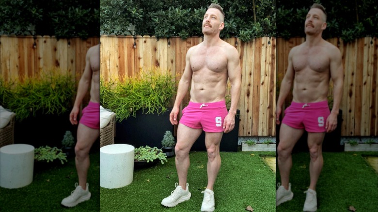 Orlando Soria in front of a wooden fence outside, wearing small pink shorts and white gym shoes and nothing else but lots of muscles.