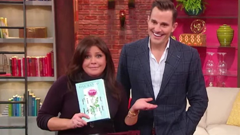 Rachael Ray holding up a copy of Bill Rancic's romance novel