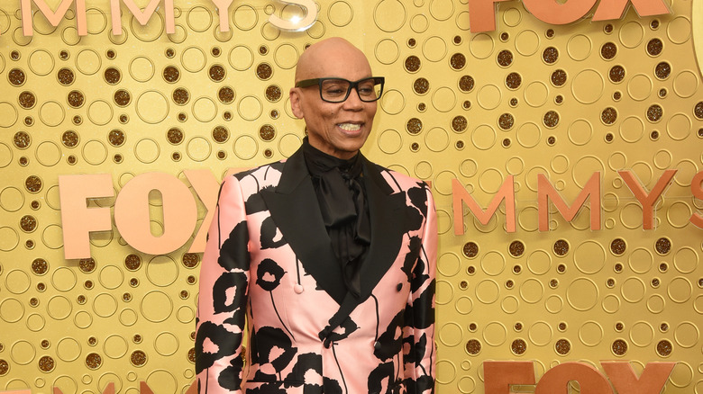 A smiling RuPaul in a black and pink suit with a yellow background 