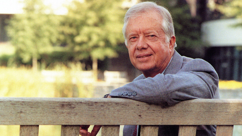 Former president Jimmy Carter leaning on a bench outside, smiling just a bit at the camera.