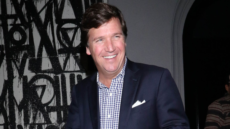 Tucker Carlson in a checked shirt and blue blazer, smiling, looking to the side of the camera