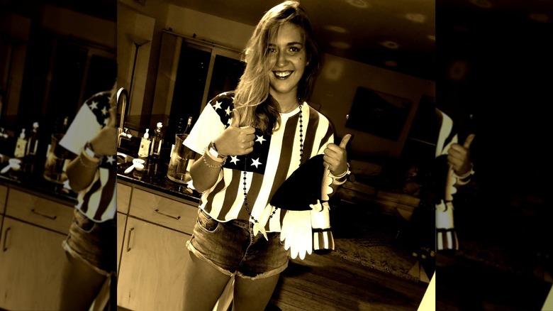 Black and white photo of Hopie Carlson wearing a flag t-shirt, jean shorts, and raising two thumbs up while smiling at camera.