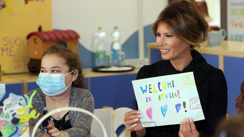 Melania Trump sitting next to child in mask, holding sign that says "Welcome in Italia"