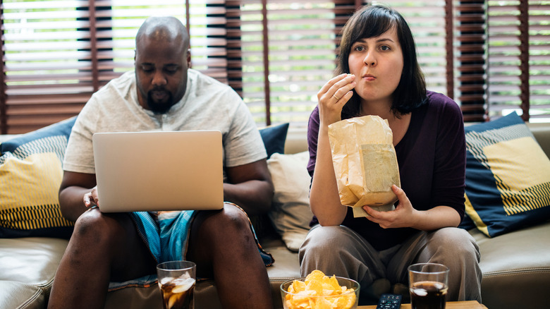 man is on computer as woman eats popcorn and watches tv