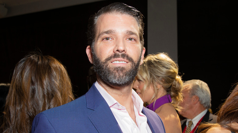 Donald Trump Jr. smiles while wearing a blue jacket and open collared shirt.