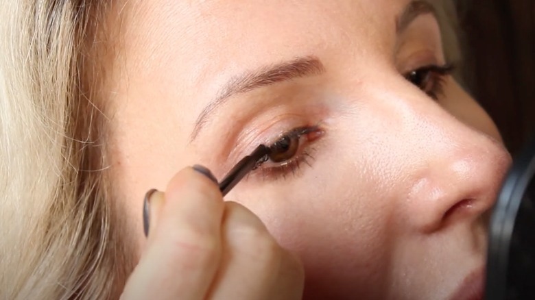 Blonde woman applies Latisse to upper lashline with black brush. 